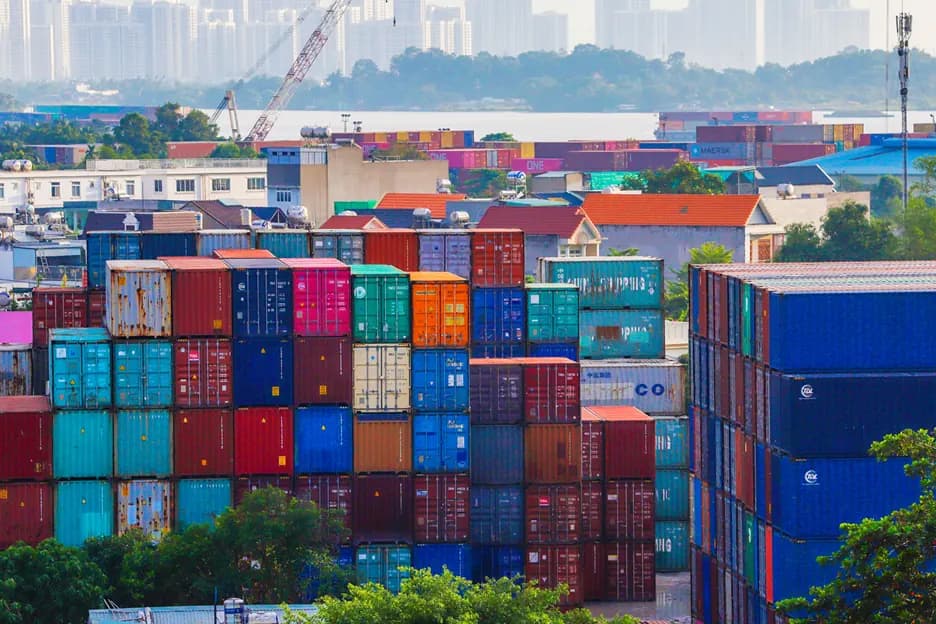 Stacked shipping containers at a port terminal, with a city skyline beyond